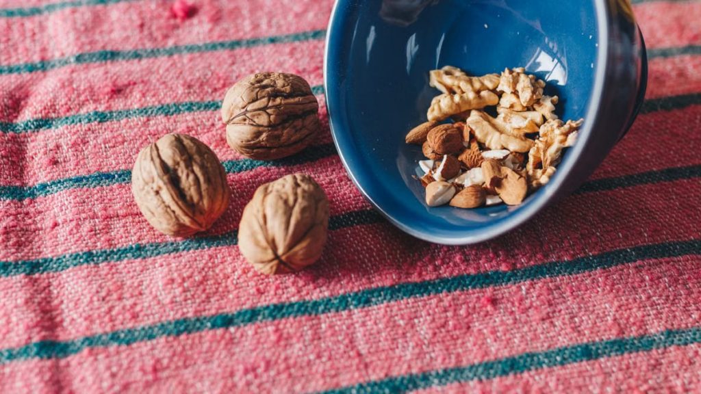 A variety of nuts, including almonds and walnuts, spilled from a blue bowl onto a pink textile surface.
